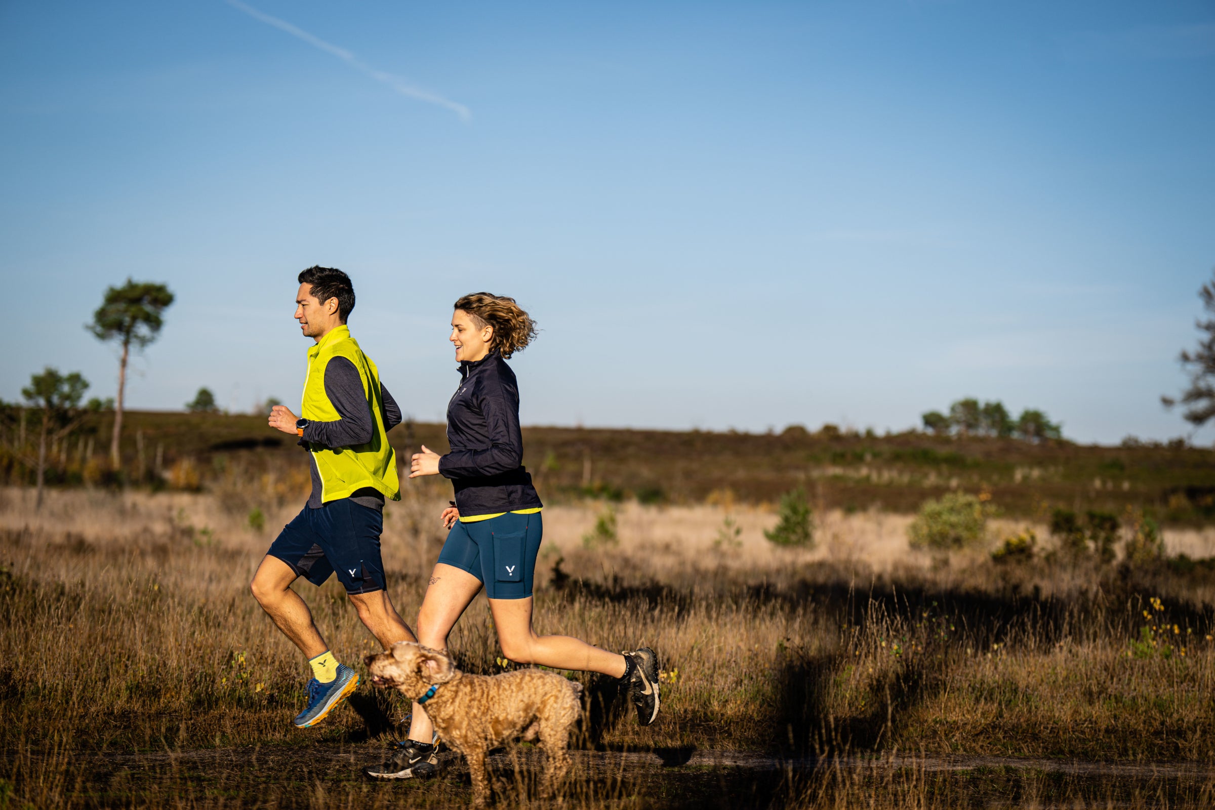 A man and a woman jogging on a heathland trail with a dog, both wearing Flyte sportswear