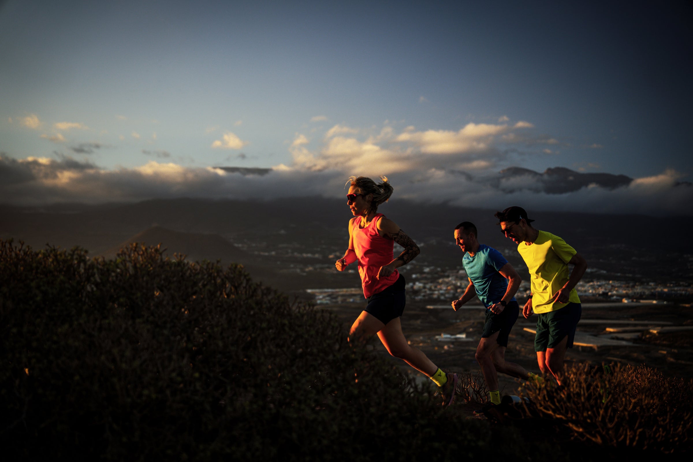 Trail runners moving uphill at sunset with mountains and clouds in the background