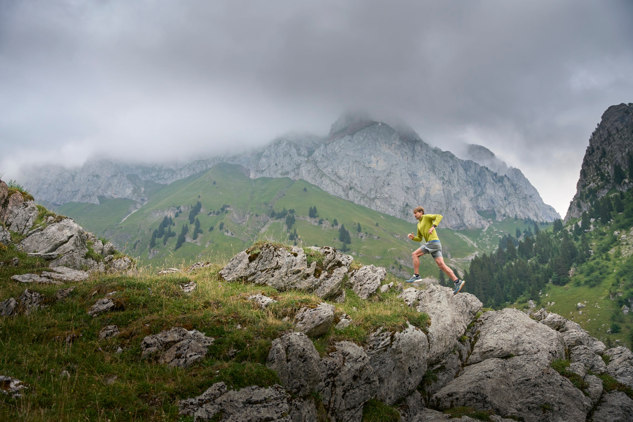 A trail runner in a bright yellow top and blue shorts running along a rocky grassy mountain ridge
